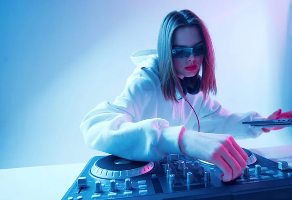 Cool young girl DJ mixes music on a mixing console and laptop, in stylish clothes, glasses on a neon background.
