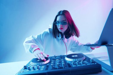 Havalı genç kız DJ karışık konsol ve dizüstü bilgisayarda müziği karıştırıyor, şık kıyafetler içinde, neon arka planda gözlük takıyor..