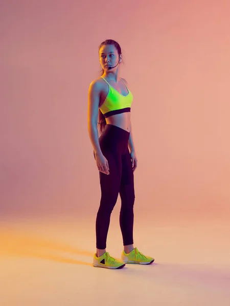 Linda chica instructor de fitness con auriculares de micrófono posando en un entrenamiento en línea, sobre un fondo de neón . — Foto de Stock