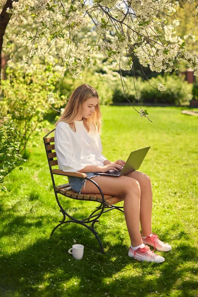 Egy aranyos tini lány sms-ezik a közösségi oldalakon egy laptopon, ül egy széken az udvaron, a gyepen, a kertben. — Stock Fotó