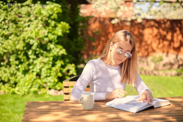 Egy aranyos fiatal lány könyvet olvas, miközben egy fa asztalnál ül egy csésze kávéval a hátsó udvaron.. — Stock Fotó