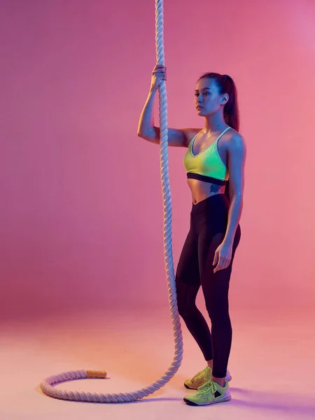 Uma linda garota de construção atlética com uma corda esportiva, após o treinamento, posa em um fundo de néon brilhante . — Fotografia de Stock