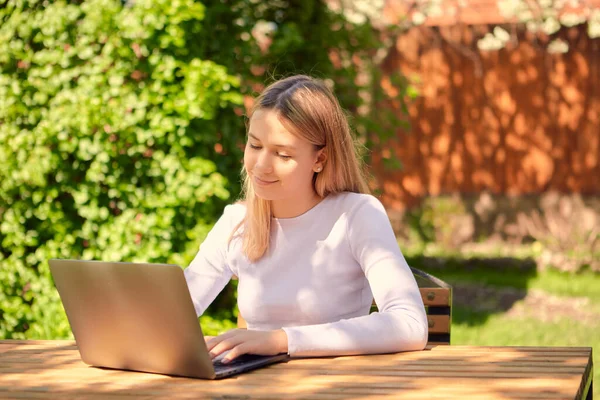 Egy aranyos fiatal lány tanul online egy laptopon, miközben ül egy fa asztal a hátsó kertben egy ház. — Stock Fotó