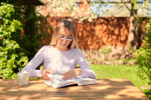 Egy aranyos fiatal lány könyvet olvas, miközben egy fa asztalnál ül egy csésze kávéval a hátsó udvaron.. — Stock Fotó