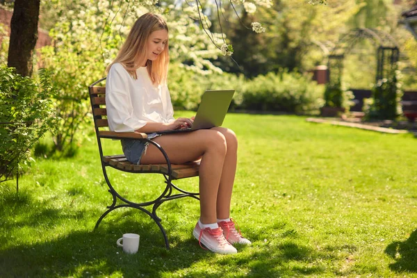 Egy aranyos tini lány sms-ezik a közösségi oldalakon egy laptopon, ül egy széken az udvaron, a gyepen, a kertben. — Stock Fotó