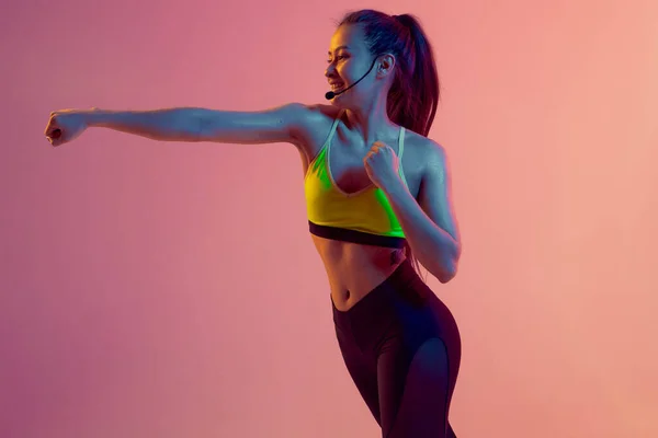 Cute girl fitness instructor teaches Boxing or body combat online training remotely on a bright neon background.