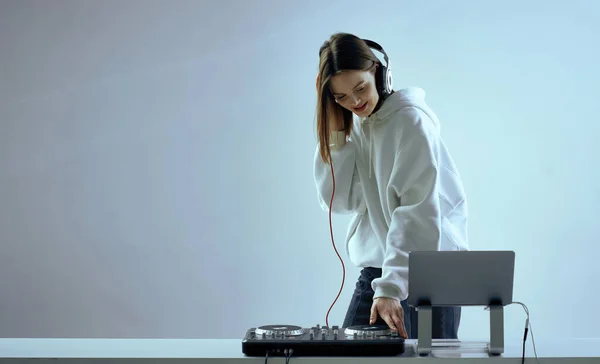 Cool mladá dívka DJ míchá hudbu na mixážní konzoli a notebook, ve stylovém oblečení, na bílém modrém pozadí. — Stock fotografie