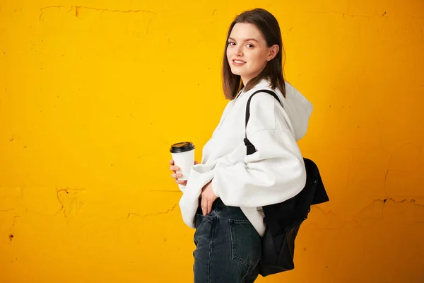 Joven chica bonita en una sudadera con capucha blanca con una mochila y una taza de café desechable posa contra una pared de hormigón amarillo . —  Fotos de Stock