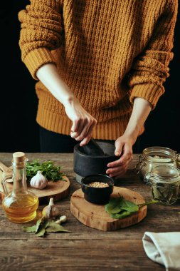 Genç ve güzel bir kız yemek pişirirken havanda baharat öğütür. Taze malzemeler ahşap bir masaya serilir..