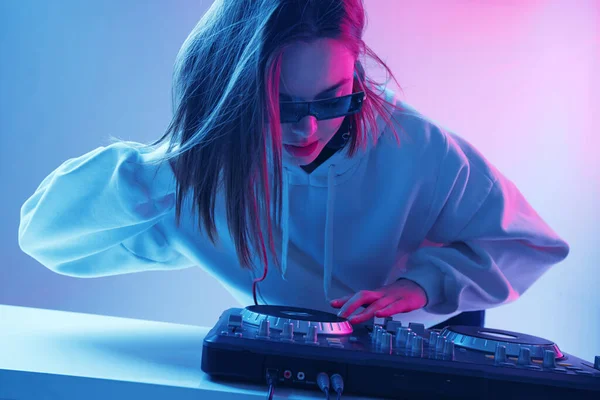 Cool jong meisje DJ mixt muziek op een mengpaneel en hoofdtelefoon, in stijlvolle kleding, bril op een neon achtergrond. — Stockfoto