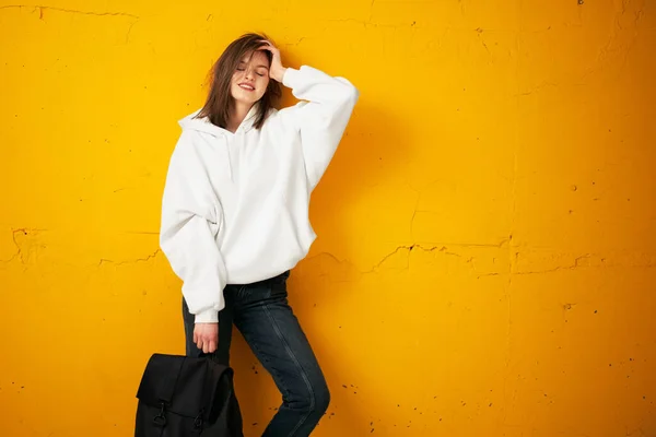 Portré egy fiatal csinos lány diák fehér kapucnis pulcsi, alkalmi ruhák, egy zsák, a beton narancs fal. — Stock Fotó