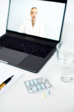Bilgisayarın arka planında bir bardak suyla birlikte tabletlerle ve tedavi için ilaçlarla su toplaması ve doktorla görüntülü konuşma.