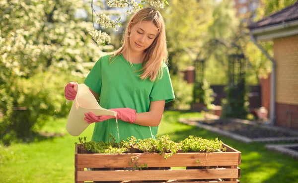 Linda muchacha adolescente feliz regando flores, en una olla de madera, de una regadera, en su jardín casero . — Foto de Stock