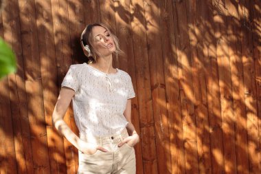 Kablosuz kulaklıklı güzel bir kızın portresi, güneşli bir günde arka bahçede ahşap bir çitin karşısında duruyor..