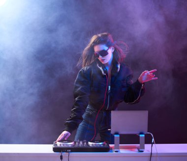 Gözlüklü şık bir DJ ve bir bombacı, dumandaki neon ışıkta dans eden bir partide müziği karıştırıyor. .