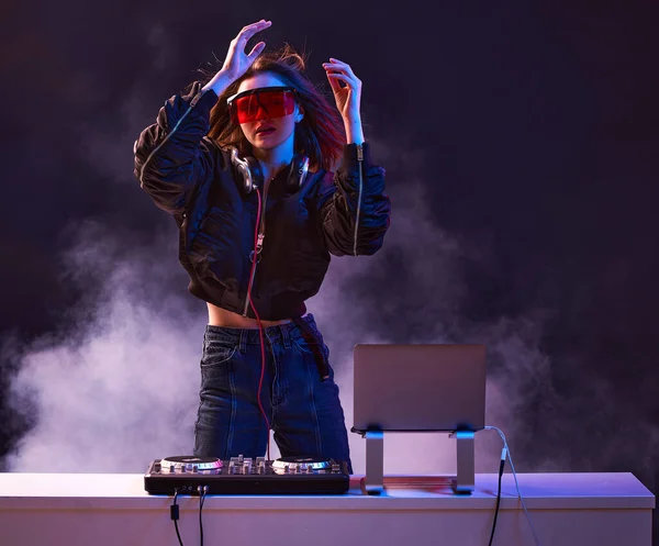 Chica con estilo DJ en gafas auriculares y un bombardero mezcla música bailando en una fiesta en la luz de neón en el humo  . — Foto de Stock