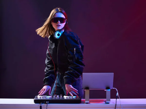 Стильна ді-джея дівчина з навушниками та окулярами на куртці бомбардувальника змішує музику на вечірці та неонових вогнях . — стокове фото