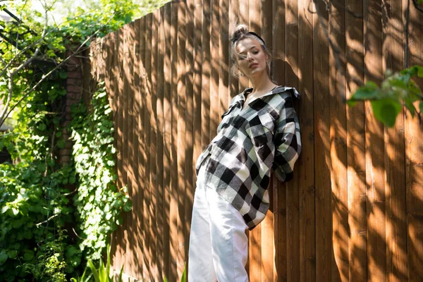 Retrato de una chica bonita con una camisa a cuadros y pañuelo de pie contra una valla de madera en el patio trasero en un día soleado . —  Fotos de Stock