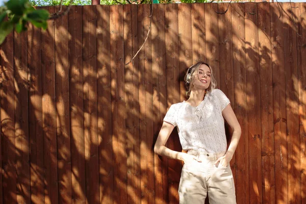 Retrato de una chica bonita con auriculares inalámbricos, de pie contra una valla de madera en el patio trasero en un día soleado . —  Fotos de Stock