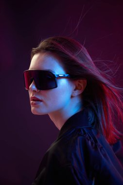 Siyah bombardıman ceketli, koyu bir arka planda güneş gözlüklü, neon ışıklı havalı bir kızın portresi..
