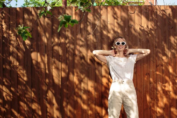 Retrato de una chica bonita con gafas y un pañuelo en la cabeza de pie contra una valla de madera en el patio trasero en un día soleado . —  Fotos de Stock