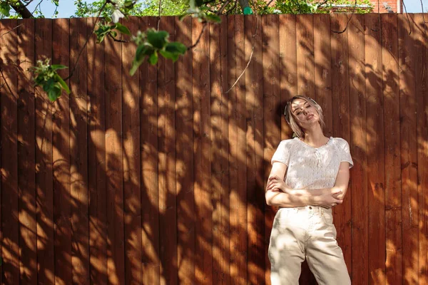 Retrato de una chica bonita con auriculares inalámbricos, de pie contra una valla de madera en el patio trasero en un día soleado . —  Fotos de Stock