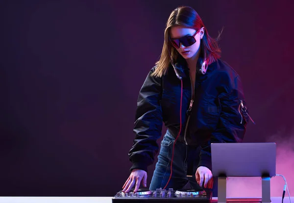 Stílusos DJ lány fejhallgatóval és szemüveggel a bombázó kabát keveri a zenét a párt és a neon fények. — Stock Fotó
