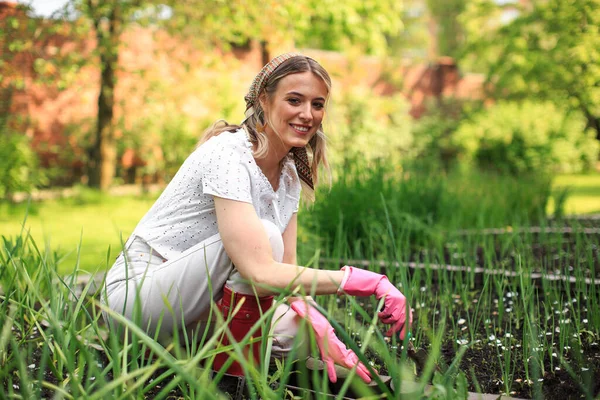 Seorang gadis muda cantik dengan selendang dan sarung tangan karet menanam bawang di kebun belakang rumahnya pada hari musim panas yang cerah. — Stok Foto