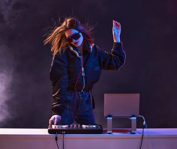 Stylish girl DJ in glasses headphones and a bomber mixes music dancing at a party in the neon light in the smoke .