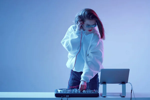 Cool mladá dívka DJ míchá hudbu na mixážní konzoli a notebook, ve stylovém oblečení, brýle na neonovém pozadí. — Stock fotografie