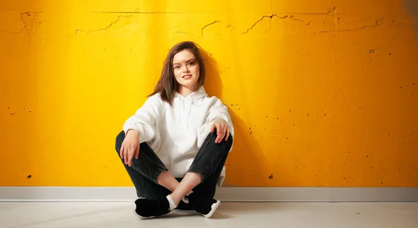 Junge hübsch lächelnde Studentin in weißem Kapuzenpulli posiert auf dem Boden vor einer gelben Betonwand. — Stockfoto