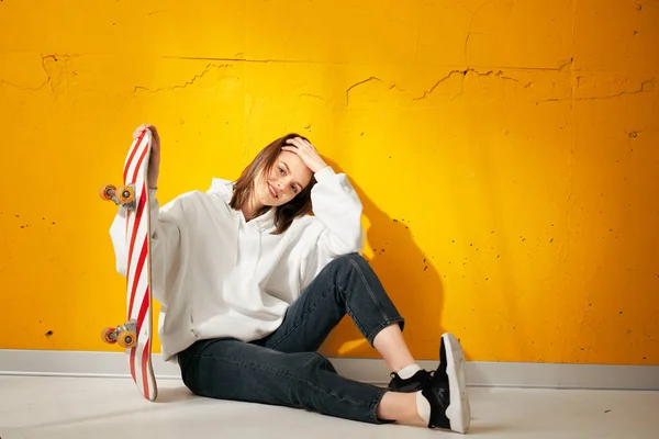 Joven chica sonriente bastante feliz en una sudadera con capucha blanca con un monopatín posa contra una pared de hormigón amarillo . —  Fotos de Stock