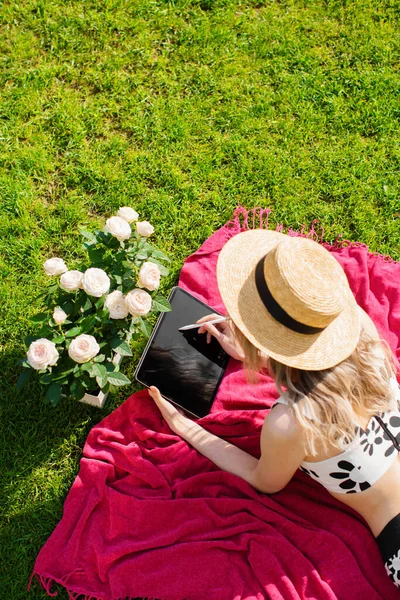 Young woman Illustrator in hat draws on tablet on green lawn with roses, photo for advertising or blog