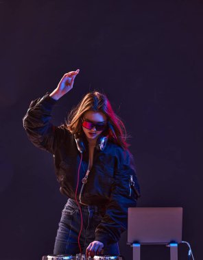 Şık bir DJ kız, kulaklıkları ve gözlükleriyle bombacı ceketinde müziği ve neon ışıkları karıştırıyor..