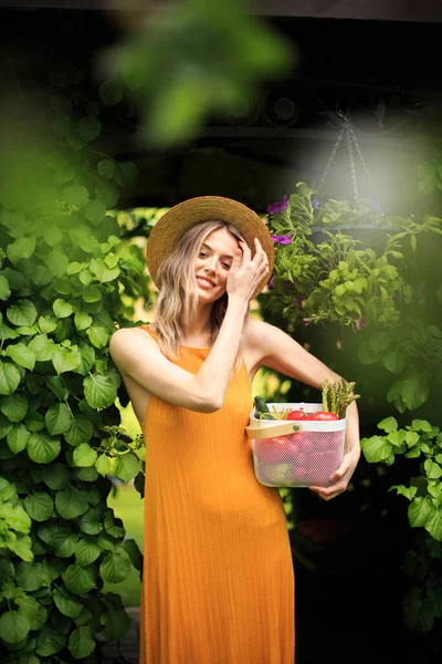 Gadis tersenyum muda dengan topi di kebun hijau musim panas dengan keranjang sayuran segar, fotografi untuk blog dan iklan — Stok Foto