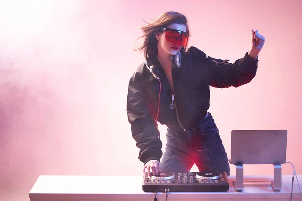 Κομψό κορίτσι DJ με γυαλιά ακουστικά και ένα βομβαρδιστικό αναμιγνύει μουσική χορεύοντας σε ένα πάρτι στο νέον φως στον καπνό . — Φωτογραφία Αρχείου