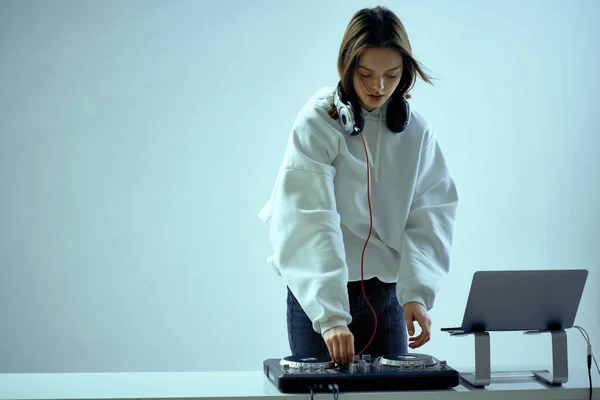 Cool mladá dívka DJ míchá hudbu na mixážní konzoli a notebook, ve stylovém oblečení, na bílém modrém pozadí. — Stock fotografie
