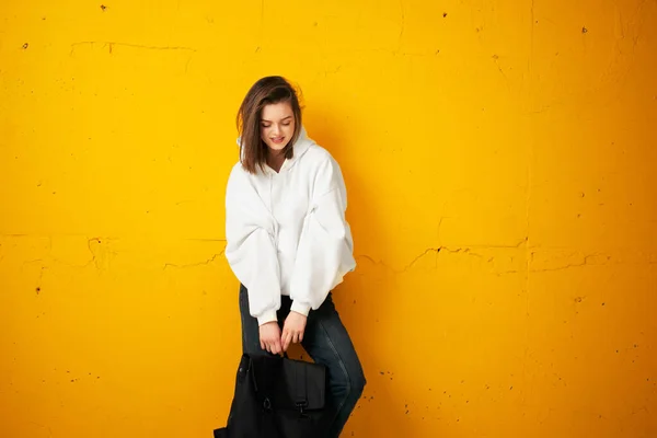 一个穿着白色连帽衫，便衣，提着袋子，靠着混凝土橙色墙壁的年轻漂亮女学生的画像. — 图库照片