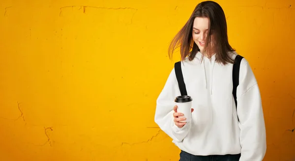 Fiatal csinos lány fehér kapucnis pulcsiban, hátizsákkal és egy eldobható csésze kávéval egy sárga betonfalnak pózol.. — Stock Fotó