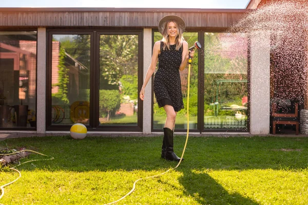 Joven mujer sonriente en sombrero y vestido negro riego césped con manguera de jardín en el patio trasero, foto para el blog o anuncio —  Fotos de Stock