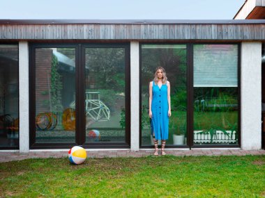 Yazlık elbiseli güzel bir kız arka bahçedeki havuz penceresinin arkasında şişme bir topla poz veriyor..