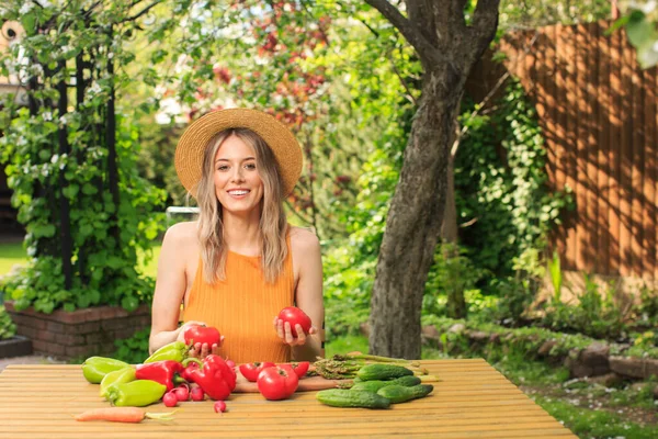 Seorang gadis cantik mengenakan topi duduk di meja kayu dengan sayuran segar tumbuh di kebunnya pada musim panas. — Stok Foto