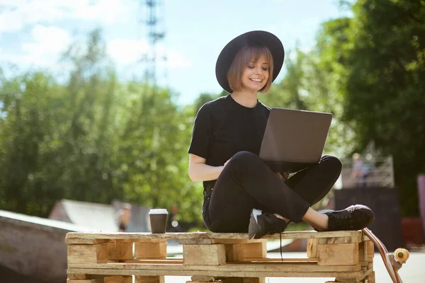 Creative young smiling woman blogger work on laptop on content sitting on wooden pallet with cup of coffee in skatepark