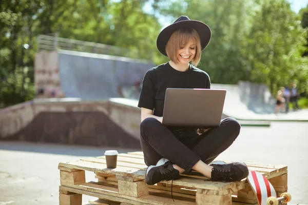 Creative young happy girl blogger work on laptop on content sitting on wooden pallet with cup and skateboard in park