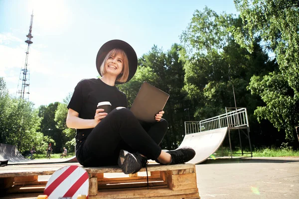 Kreatív fiatal boldog lány blogger munka laptop tartalmat ül fa raklap csésze és gördeszka a parkban — Stock Fotó
