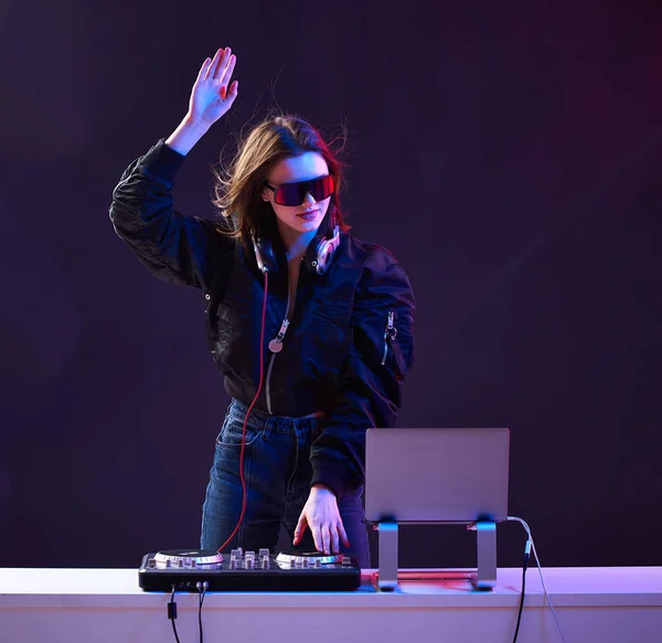 Stylish DJ girl with headphones and glasses on the bomber jacket mixes the music at the party and neon lights.
