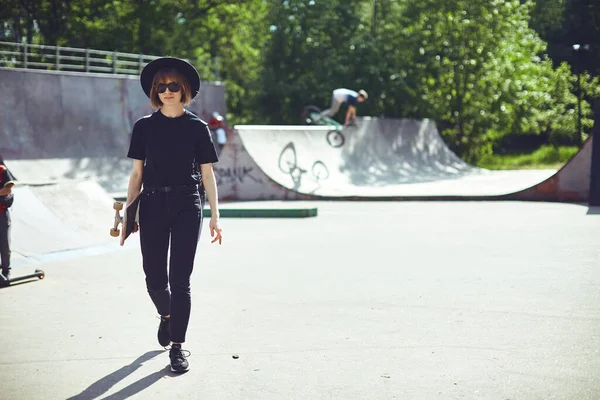 Ziemlich flippiges Teenager-Mädchen mit Longboard, das in Sonnenbrille, Hut und schwarzer Kleidung im Skatepark spazieren geht, für Werbung oder Blog — Stockfoto