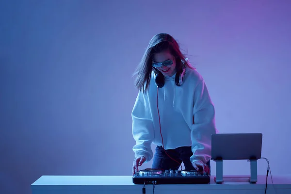 Cool joven DJ mezcla música en una consola de mezcla y un ordenador portátil, con ropa elegante, gafas sobre un fondo de neón . — Foto de Stock