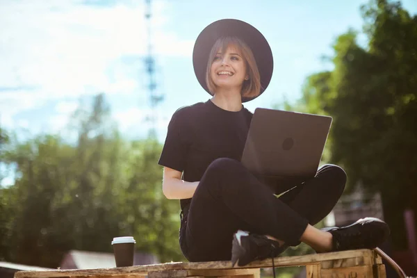 Creative young smiling girl blogger work on laptop on content sitting with a cup of coffee to go in park