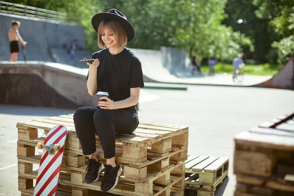 Mosolygós lány chat telefonon ül a fa raklapon a parkban gördeszka és csésze kávé menni, fotó a reklám vagy blog — Stock Fotó